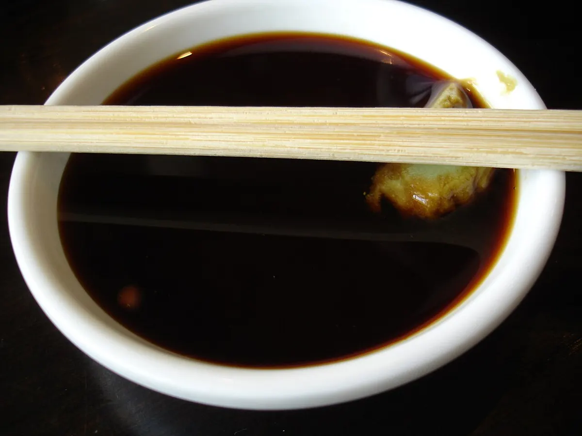 A small bowl of soy sauce with a blob of green wasabi paste on the side and chop sticks laid across the top.